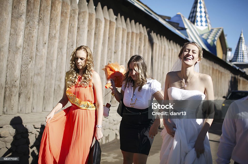 Bride and her bridesmaid walking with friends Laughing happy bride and her bridesmaid walking with their friends along a road outdoors alongside a wooden palisade Adult Stock Photo