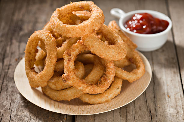 Onion Rings A high angle close up shot of a pile of Onion  Rings stacked on a "recycled" bamboo plate and a small container of ketchup in the foreground. Shot on an  old grungy wooden picnic table. fried onion rings stock pictures, royalty-free photos & images