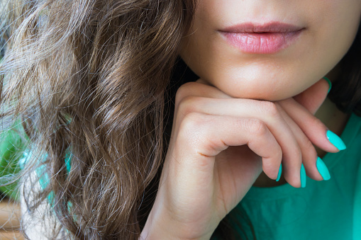 young beautiful girl with brown shiny hair holds a hand with a turquoise nail polish near the chin, lips dyed red lipstick. the image is dominated by green, the model's face is not visible