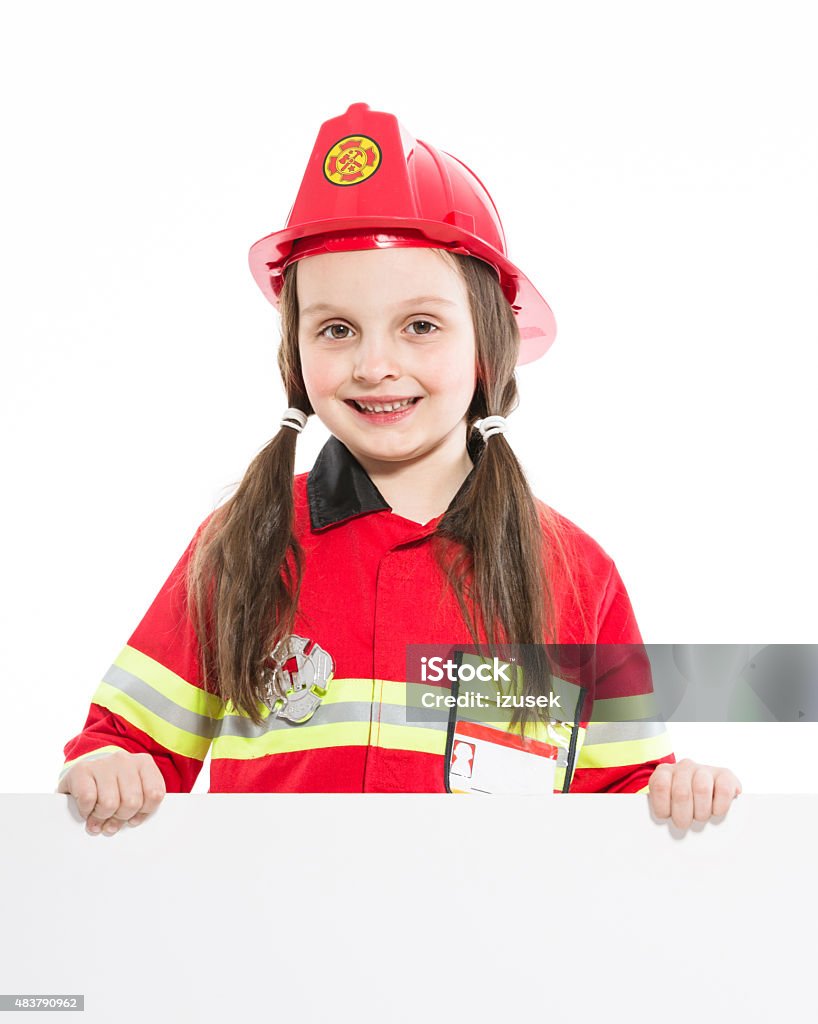 Happy firefighter girl holding whiteboard Portrait of little girl dressed as a firefighter holding whiteboard in hands and smiling at the camera. Studio shot, isolated on white. Firefighter's Helmet Stock Photo