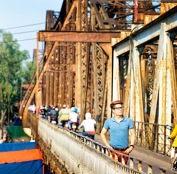 Stylish young Vietnamese man enjoying a walk on Long Bien Bridge in Hanoi, Vietnam on a warm Spring evening, while people cross on motorbike in the background. He is looking away pensively, and is wearing a blue polo shirt. He sports blonde highlights in his hair. Square frame.