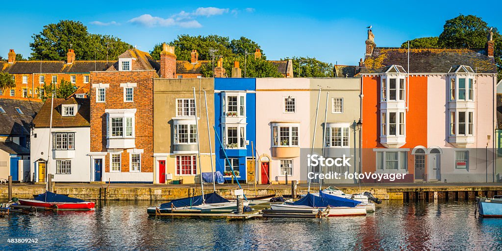 Colorful homes quaint cottages in sunny fishing village harbor panorama Colourful cottages and narrow higgledy-piggledy homes reflecting in the tranquil harbour of a quaint fishing port under the blue summer skies of Weymouth, Dorset, UK. ProPhoto RGB profile for maximum color fidelity and gamut. Weymouth - Dorset Stock Photo