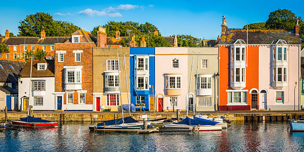 Colorful homes quaint cottages in sunny fishing village harbor panorama Colourful cottages and narrow higgledy-piggledy homes reflecting in the tranquil harbour of a quaint fishing port under the blue summer skies of Weymouth, Dorset, UK. ProPhoto RGB profile for maximum color fidelity and gamut. dorset england stock pictures, royalty-free photos & images