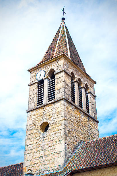 Vertical composition color photography of old french stone church steeple close-up, shot on cloudy sky background in summer season. Shot without people, in Benonce city, in Bugey mountains, in Ain, Rhone-Alpes region in France (Europe).