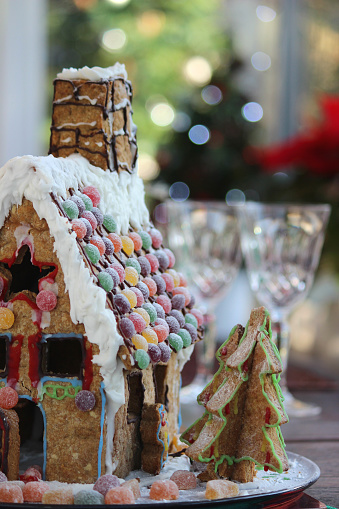 Photo showing a gingerbread house decorated with sweets and displayed on the Christmas table, which is laid ready for dinner, with wine glasses.  Roof tiles made from sweets have been used as decorations on the roof.