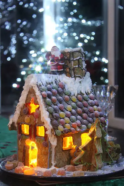 Photo showing a gingerbread house that has been photographed at night, with a Santa Claus character standing on the roof next to the chimney pot.  The house is lit with tealight candles, while the roof is decorated with jelly sweets and piped icing.