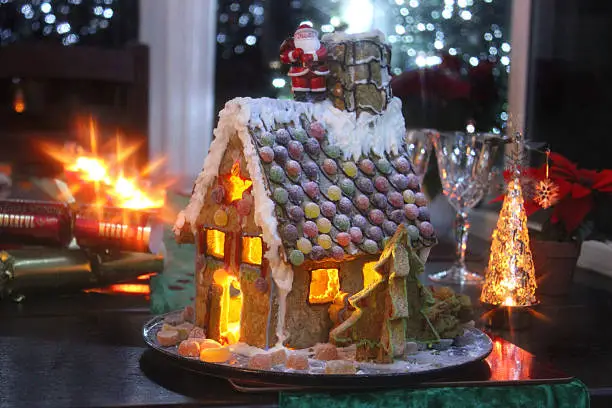 Photo showing a homemade Christmas gingerbread house that is pictured at night, when it is lit from the inside with a tealight candle.  Christmas crackers, fairy lights, a small Santa Claus and sweet decorations complete this festive scene.