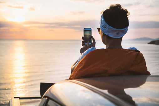 Young woman sitting on the back of pickup truck in sleeping bag taking photo of the sunset over the sea.
