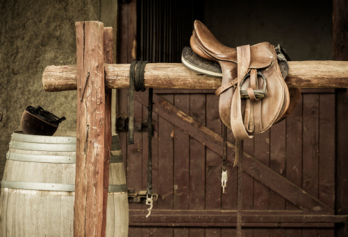 In front of a stable. A saddle, barrel and equestrian helmet.