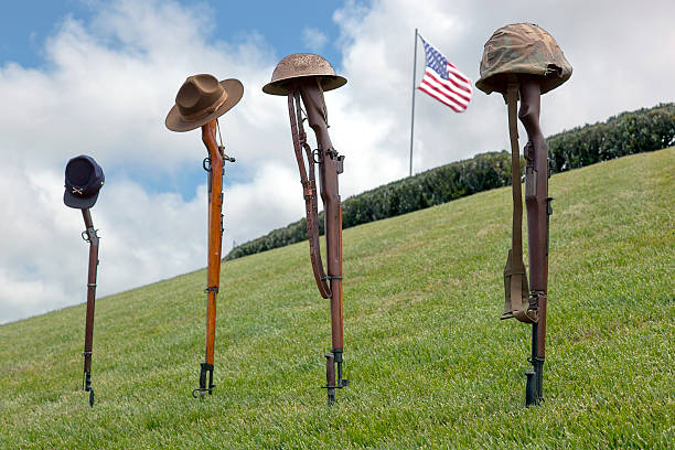 Vintage, antique rifles and soldier's hats and helmets forming Fallen Soldier Battle Crosses, American Flag behind, left to right, from eras of Civil War, Spanish American War, WWI, WWII.