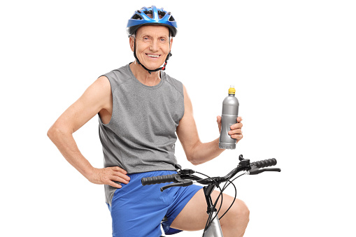 Senior man with a blue helmet holding a water bottle and posing seated on his bike isolated on white background