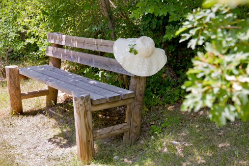 Wide brimmed summer sunhat draped over the back of a rustic wooden garden bench standing amongst green foliage in a garden in Provence, France.