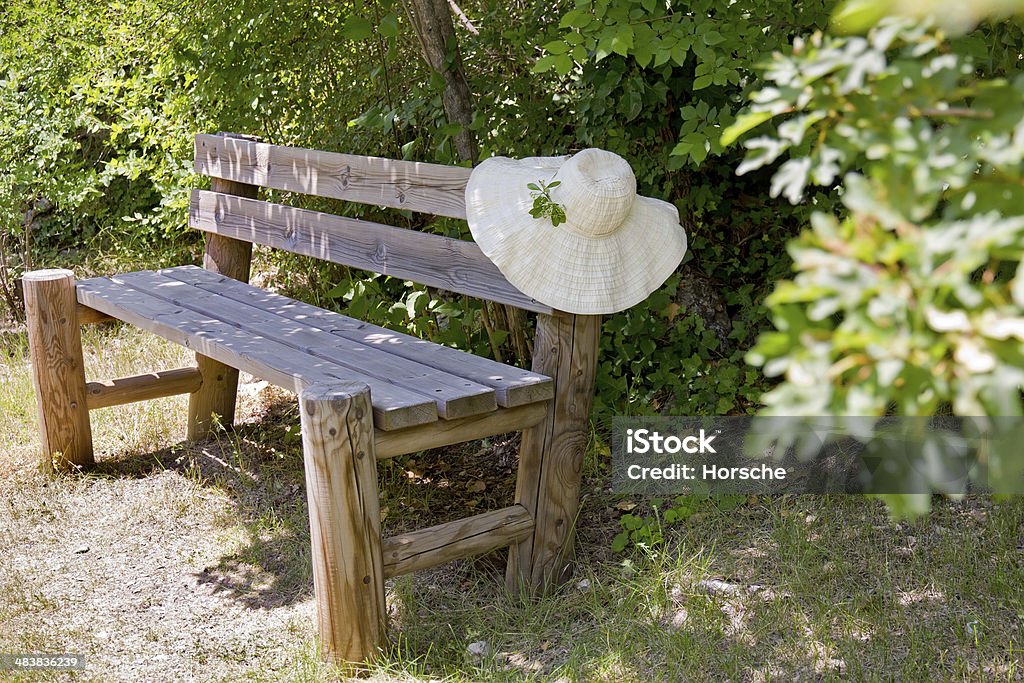 Sunhat on a wooden garden bench. Wide brimmed summer sunhat draped over the back of a rustic wooden garden bench standing amongst green foliage in a garden in Provence, France. Beauty Stock Photo