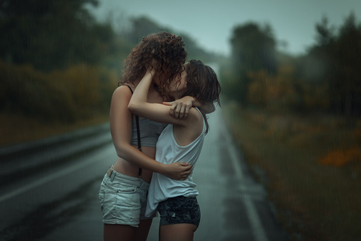 Girls standing in the rain on the street. They hug each other.