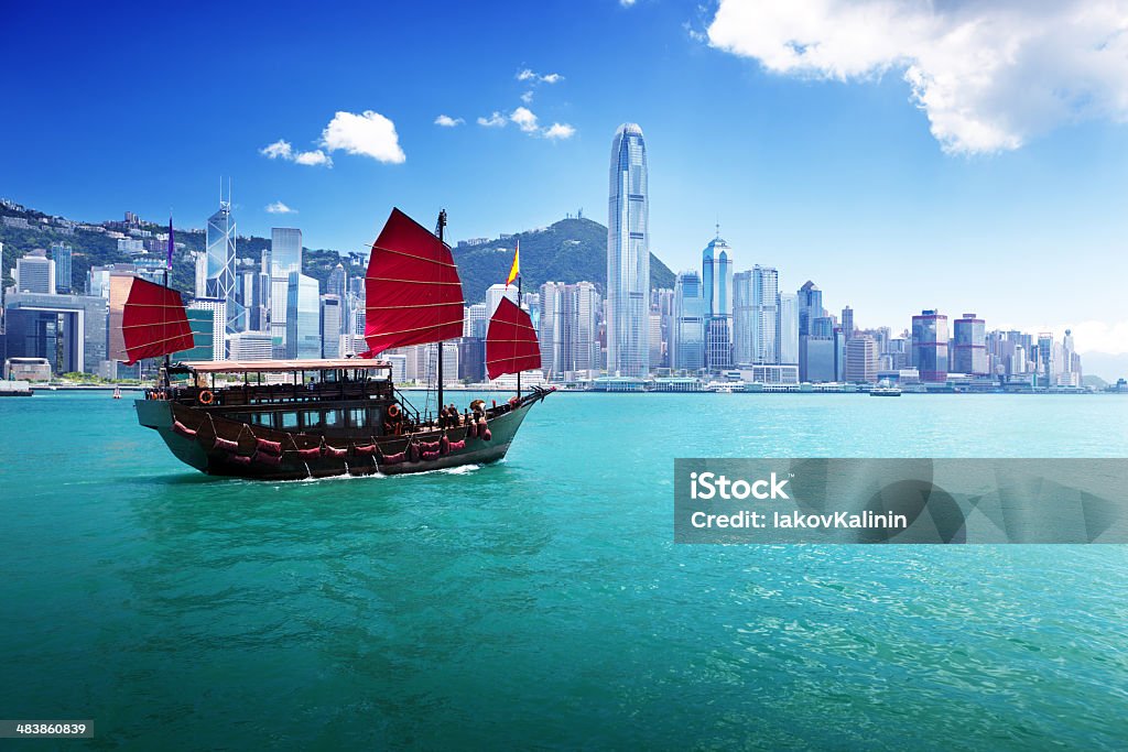 Classic sailboat in Hong Kong harbor The Hong Kong skyline from the harbor at daytime.  The water is blue-green, and a traditional Chinese junk ship with square red sails is in the water in the foreground.  The buildings in the skyline are of various heights and mostly white in front of a hillside covered with smaller buildings under a blue sky. Hong Kong Stock Photo