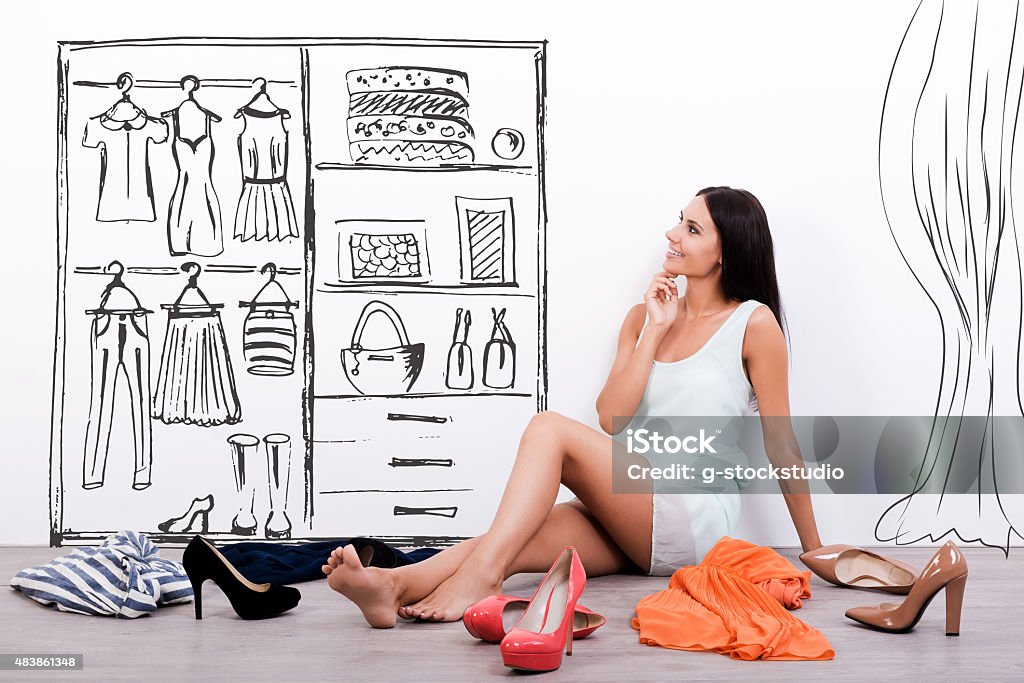 Hard choice. Thoughtful young woman in dress looking at the sketch on the wall while sitting on the floor with clothes and shoes laying around her Closet Stock Photo