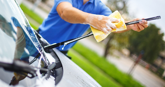 Closeup of unrecognziable adult man washing his car at a parking lot. He's using yellow spong and soap and cleaning windshield wipers. The man is wearing blue polo shirt.