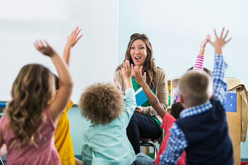 A female, Asian preschool or kindergarten teacher in the classroom, sitting in front of a group of multi-ethnic children, reading a book.  The boys and girls are raising their hands to answer a question.  There is a large, blank whiteboard in the background for copy.