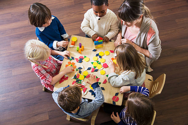 Teacher with preschoolers playing with colorful shapes An overhead view of a multiracial group of six preschoolers sitting with their teacher at a table playing with colorful blocks and geometric shapes.  The little girls and boys, 4 and 5 years old, look serious and busy putting the shapes together. toy block photos stock pictures, royalty-free photos & images