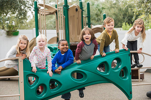A multiracial group of six children playing on a playground in preschool.  The boys and girls are climbing on the playground equipment, in a row.  Some are looking at the camera, laughing and smiling and making faces.