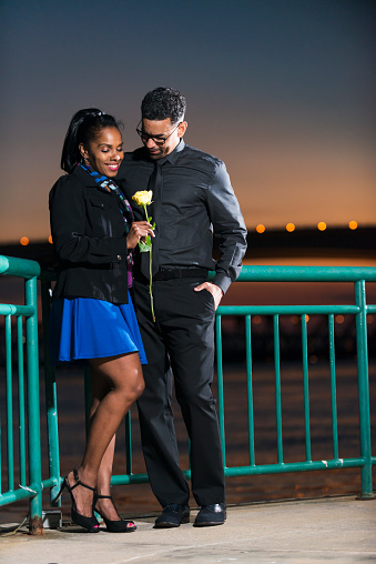 A mature, affectionate, African American couple, stylishly dressed, out in the evening on a romantic date.  They are in their 40s, standing together by the water, bridge and sky in the background.  The woman is smiling and looking down at the single, yellow rose she is holding in her hand. She is wearing a black jacket and a colorful scarf over a royal blue dress and high heels.  He is wearing dark gray pants, shirt and tie.