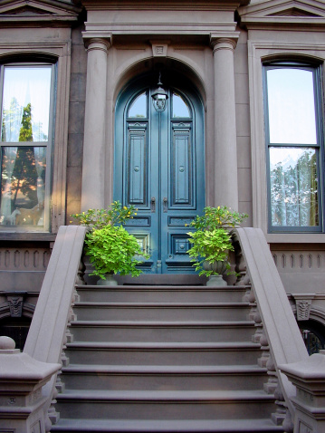 The grand entrance to a recently restored residential brownstone. Two potted plants flank each other at the top landing of the stoop. The tall, arched entry doors are painted a rustic blue. (More and similar photos are in the \