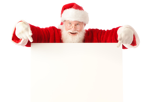 A Christmas Santa Claus dressed in a traditional Santa suit, pointing to a blank white message board, available space for adding a holiday greeting, retail advertisement, marketing message, or invitation information. Photographed in studio on white background in horizontal format.