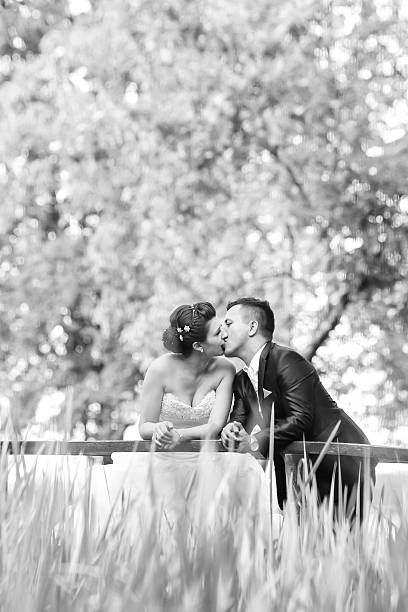 The bride and groom standing on a wooden bridge in nature, leaning on the fence and kissing.