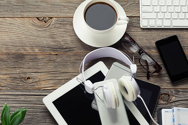 High angle shot of mobile devices and headphones. There are several smart phones and a digital tablet along with a keyboard. Devices are on a wooden table with coffee. Copy space on right.
