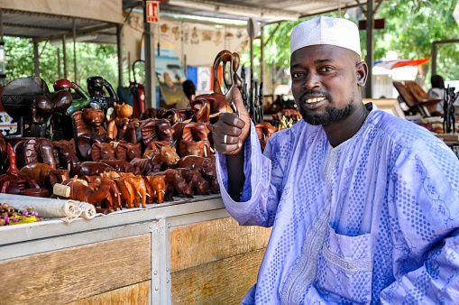 N'Djamena, Chad - October 16, 2012: African man selling in street market goods, he is happy, smiley and showing his thumb up, wearing traditional clothes. His business going well, he is offering beautiful statue of elephants from wood material.