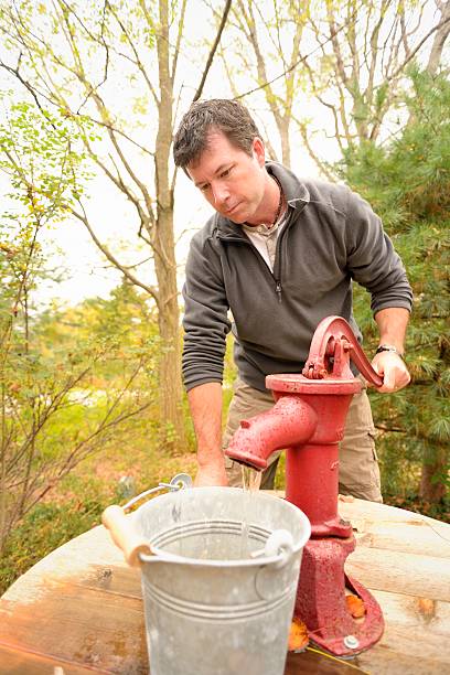 Pumping Water from a Well A casually dressed mature man using a hand pump to fill a metal bucket with water from a well. old fashioned well hand pump stock pictures, royalty-free photos & images