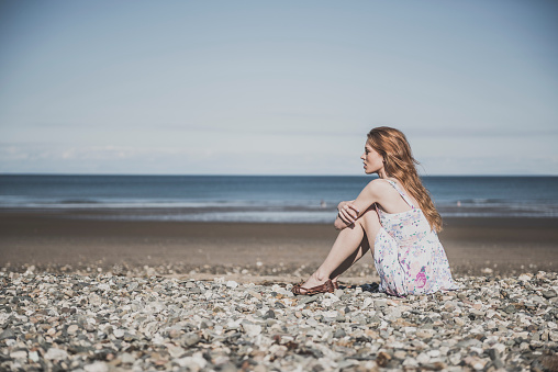 Attractive young woman with long red hair sitting thoughtfully on a pebble beach.