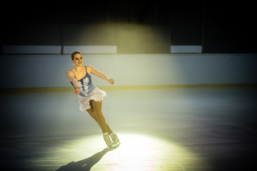 Young female figure skater performing on ice rink, arm outstretched.