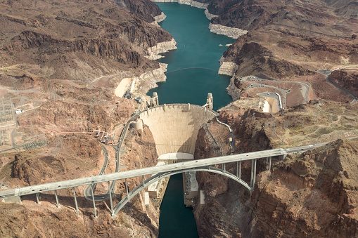 Aerial view of the Colorado River Bridge and the Hoover Dam in Nevada, Arizona, USA. There is a hydroelectric power station, a bridge and a dam wall. On either side of the bridge are rock formations that feature roadways. Popular travel destination.