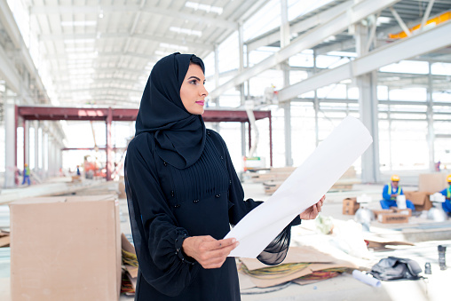 Middle Eastern businesswoman checking blueprints at construction site. She is wearing abaya, traditional Emirati clothes. We can see some construction workers in back.