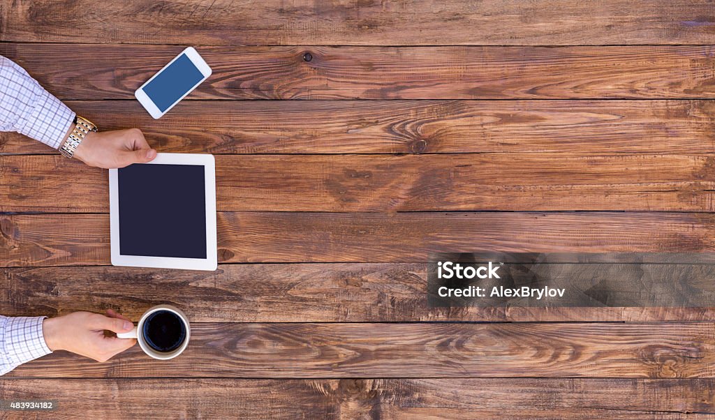 From above image of man browsing gadget empty wood desk Natural wood desk directly top view man arms holding white tablet PC coffee mug black screen cell phone casual dress shirt cuffs red light brown board plank browsing internet Wood Grain Stock Photo