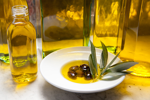 Close up view of an extra virgin olive oil bottle pouring oil on a glass bowl filled with various types of organic olives shot on rustic wooden table. Olive tree branches complete the composition. High resolution 42Mp studio digital capture taken with Sony A7rII and Sony FE 90mm f2.8 macro G OSS lens