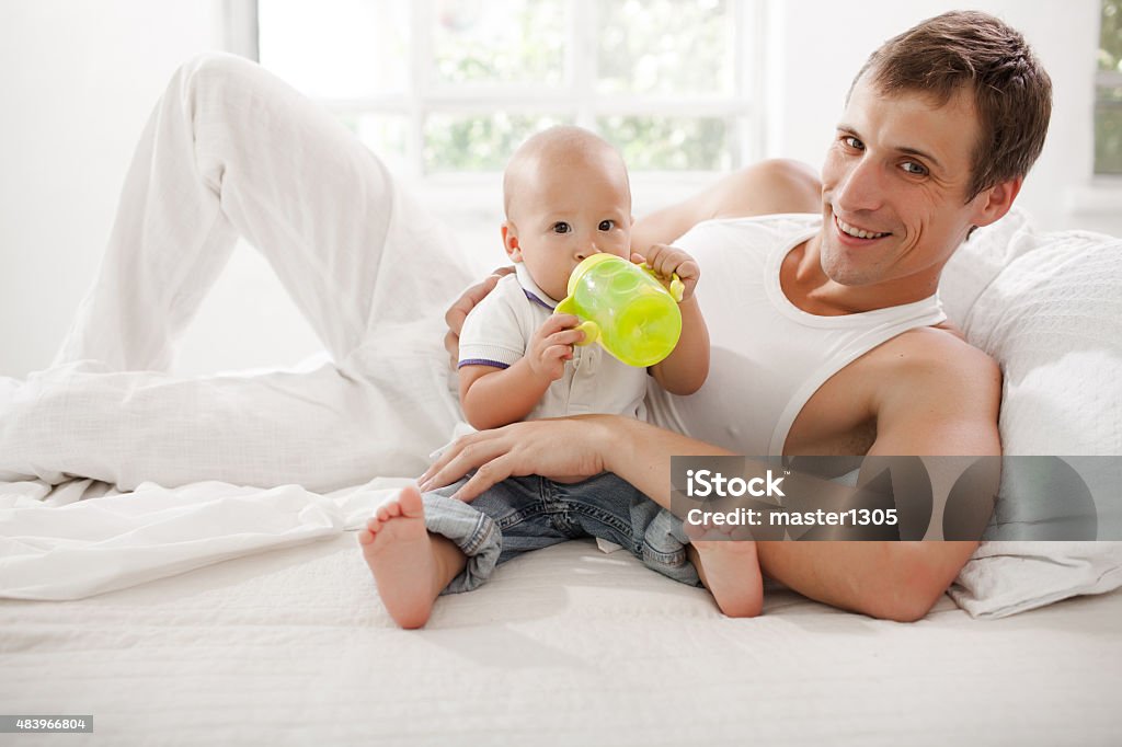 Young father with his nine months old son on the Young smiling father with his nine months old son on the bed at home on white home background. Child drinking water 2015 Stock Photo