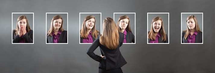 Concept photo for personality, character and emotional expression. A woman in business suit holding up a photo of herself in front of her with various range of emotional expressions exhibited in the background wall behind her. From happiness to sadness, anger to joy, apprehension to confidence. Photographed in panoramic horizontal format in studio.