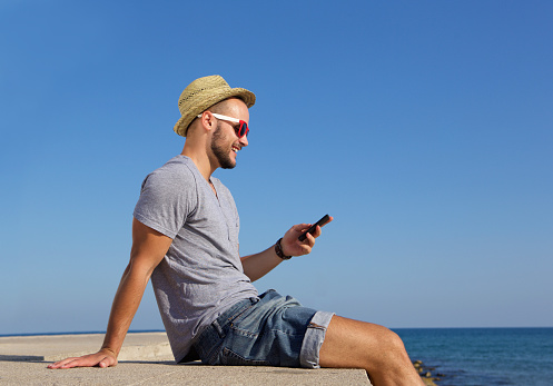 Side portrait of a happy young man sitting by the sea with mobile phone