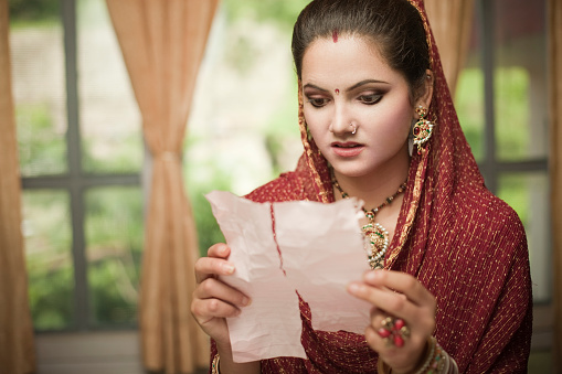 Indoor day time image of a beautiful, newly married Indian young, worried and disturbed woman from a wealthy family standing against a window background by holding two torn wrinkled pieces of a paper to which she is recombining and reading. She is in traditional Hindu dress that is Salwar Kameez, dupatta and bangles. Horizontal composition with copy space and selective focus.