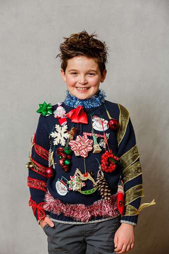 A child wearing a homemade ugly Christmas sweater using ornaments, ribbons and garland on a gray backdrop.