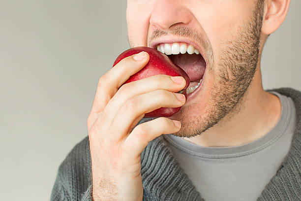 Closeup of a young man eating an apple Closeup of a young man with a beard eating an apple on a grey plain background apple bite stock pictures, royalty-free photos & images