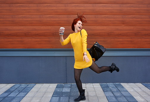 funny woman holding coffee cup and jumping, energy and fun moment.urban scene, wall in background.