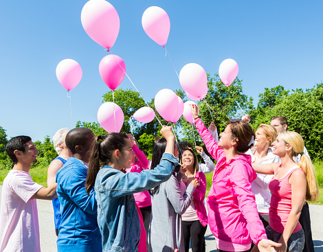 Diverse group of men and women are gathered in park on sunny summer day. They are releasing pink balloons to honor breast cancer survivors. They are wearing athletic clothing, and are in park to race for breast cancer awareness and research charity.