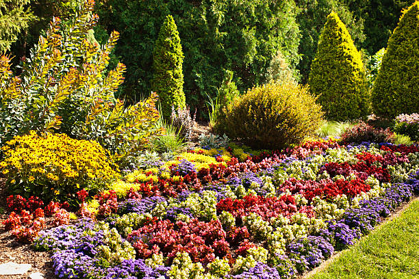 Landscaped front or back yard garden area next to a tree forest, with formal, ornamental gardens, flower beds, bushes, hedges, lawn, and rustic stone path. The tranquil summer park scenic landscape has no people and displays lush foliage of a natural outdoor shaded space for relaxation and quiet reflection.
