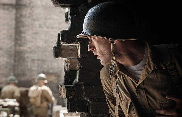 Depicted here is the rear view of a Second World War US infantryman, at rest in his dugout while in the background the tracks of armoured vehicles and tanks are visible as they move around and prepare for battle.\n\nThis image was taken at an outdoor event over the weekend of 11th-13th August 2023, where reenactors, living historians, and members of the public can come to experience what life was like during the second world war, with live demonstrations, battle reenactments, and static displays from all over the world.
