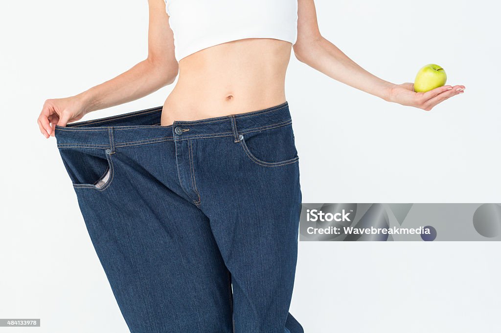 Slim woman wearing too big jeans holding an apple Slim woman wearing too big jeans holding an apple on white background 20-29 Years Stock Photo