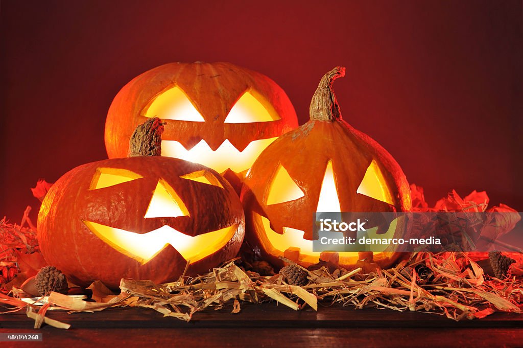 halloween pumpkins three illuminated halloween pumpkins and straw on old weathered wooden board in front of red background 2015 Stock Photo