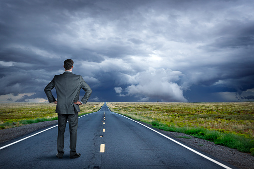 A businessman stands with his hands on his hips as he looks down a long rural road towards a fast approaching storm.